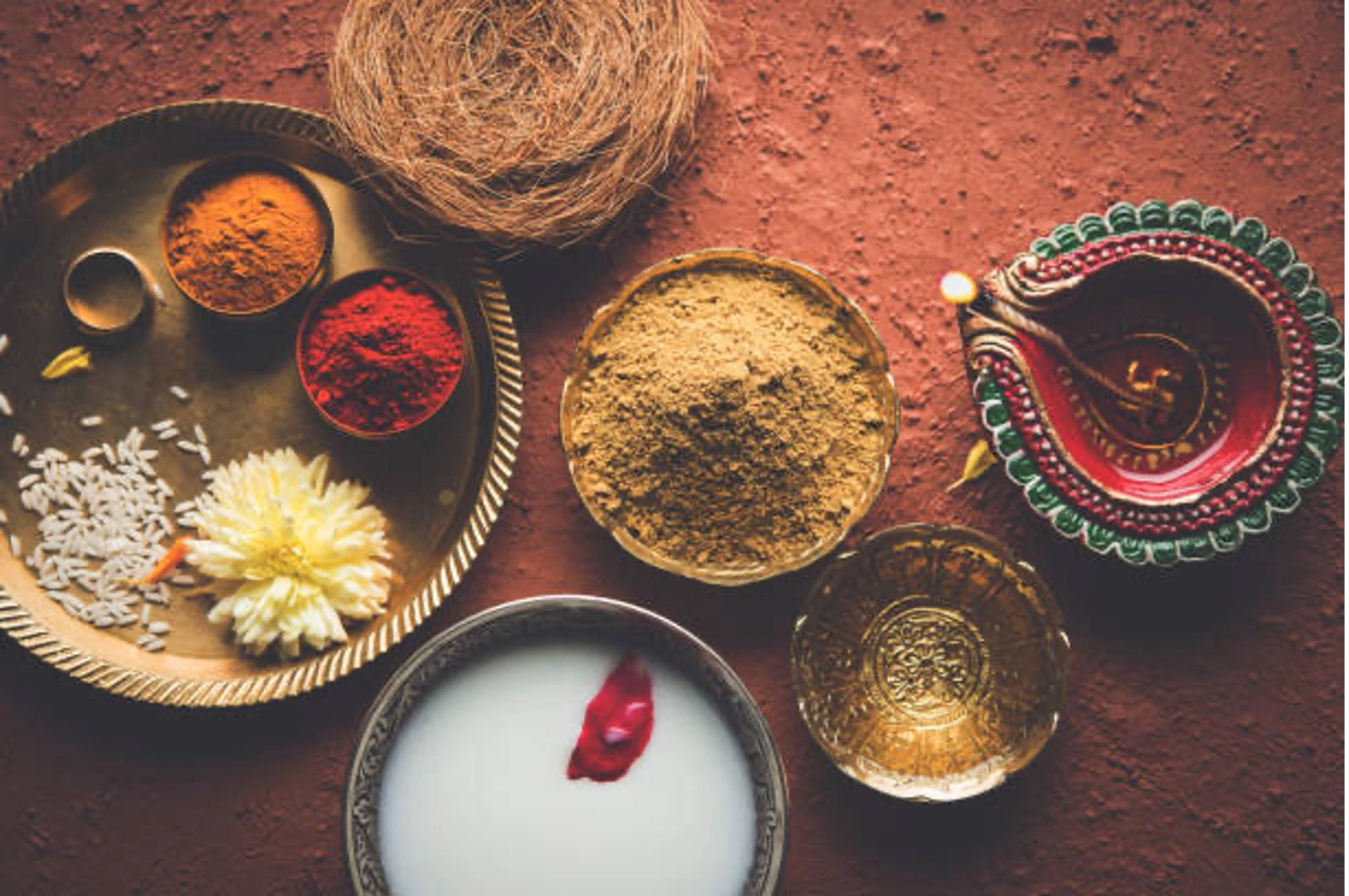 Ayur Picture 1 - Dr. Gayatri Damodaran | Ayurveda Doctor In Mira Bhayander, Mumbai Top‑down view of Hindu puja items: bowls of orange and red powders, white rice scattered on a metal tray, a coiled coconut fibre, and a decorative red‑green bowl on a reddish surface.